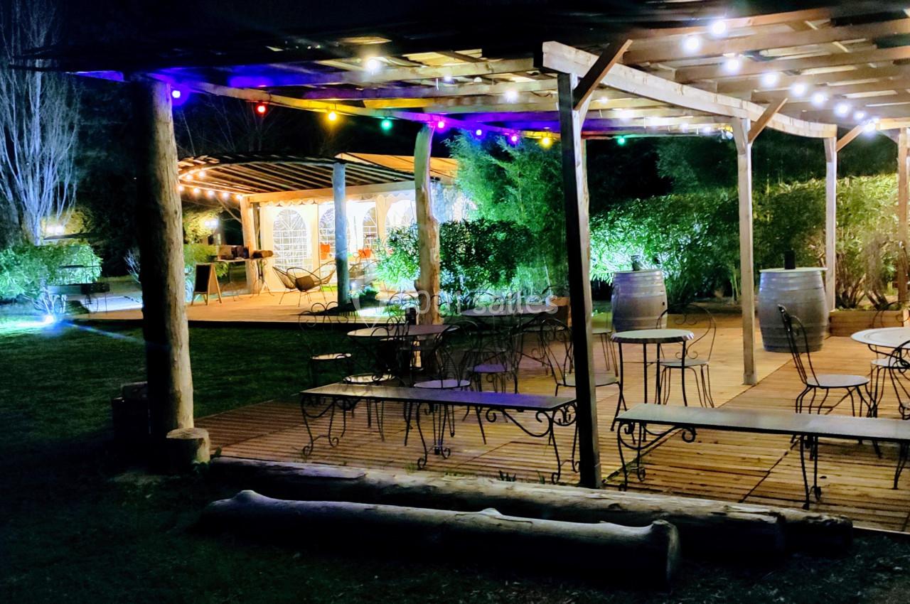 Terrasse en bois éclairée par des guirlandes lumineuses colorées, entourée de végétation, avec tables et chaises disposées.