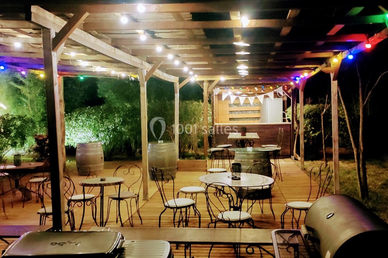 Terrasse en bois éclairée par des guirlandes lumineuses, avec tables, chaises et bar en arrière-plan, entourée de verdure.