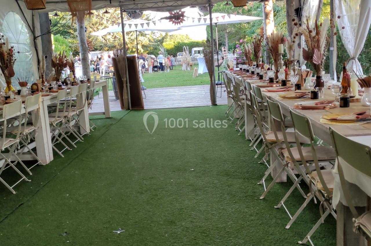Tables décorées pour un événement sous une tente, avec chaises blanches et allée centrale sur gazon artificiel.