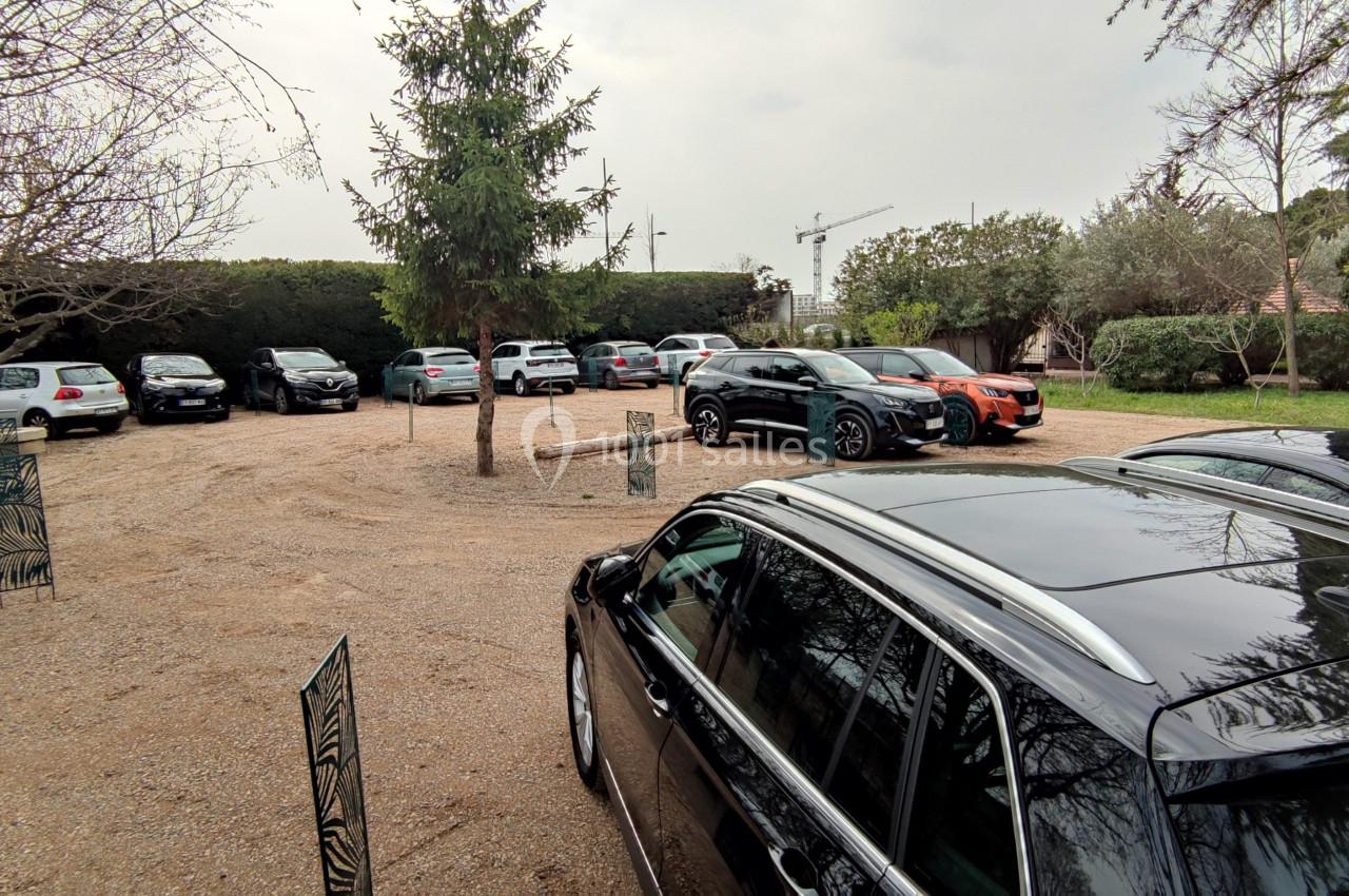 Parking extérieur avec plusieurs voitures stationnées sur un sol en gravier, entouré d'arbres et de verdure.
