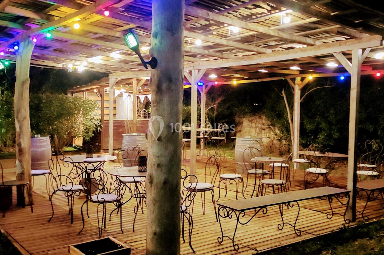 Terrasse en bois éclairée par des guirlandes lumineuses colorées, avec tables et chaises en métal, entourée de verdure.