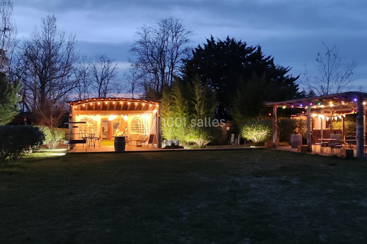 Deux pavillons éclairés dans un jardin au crépuscule, entourés d'arbres et de buissons.