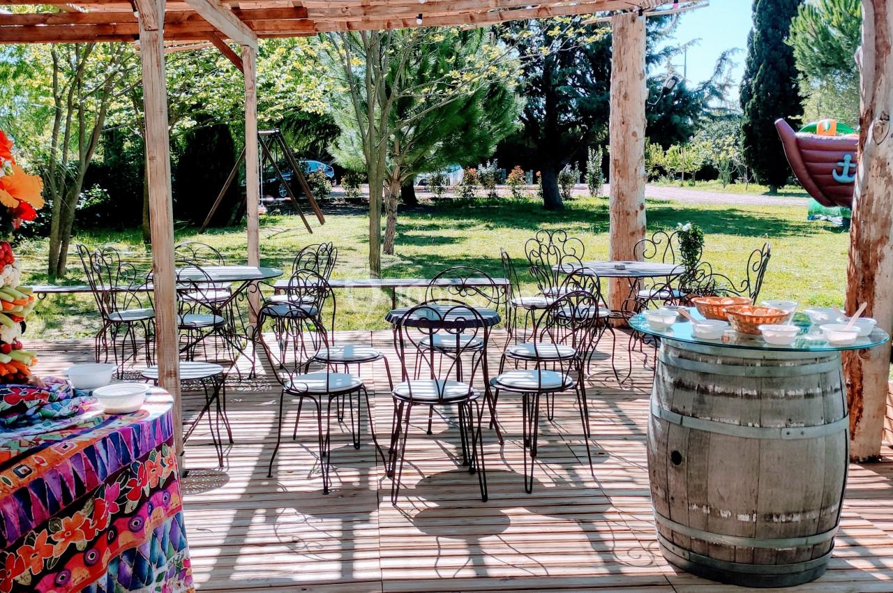 Terrasse en bois avec tables et chaises en fer forgé, décorée de fleurs, donnant sur un jardin avec structures gonflables.