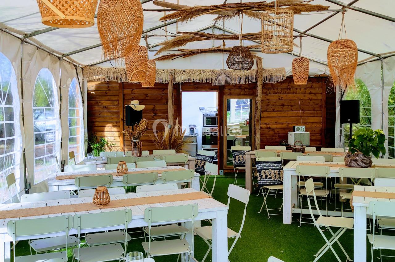 Intérieur d'une tente aménagée avec des tables et chaises blanches, décorée de luminaires et éléments en osier.