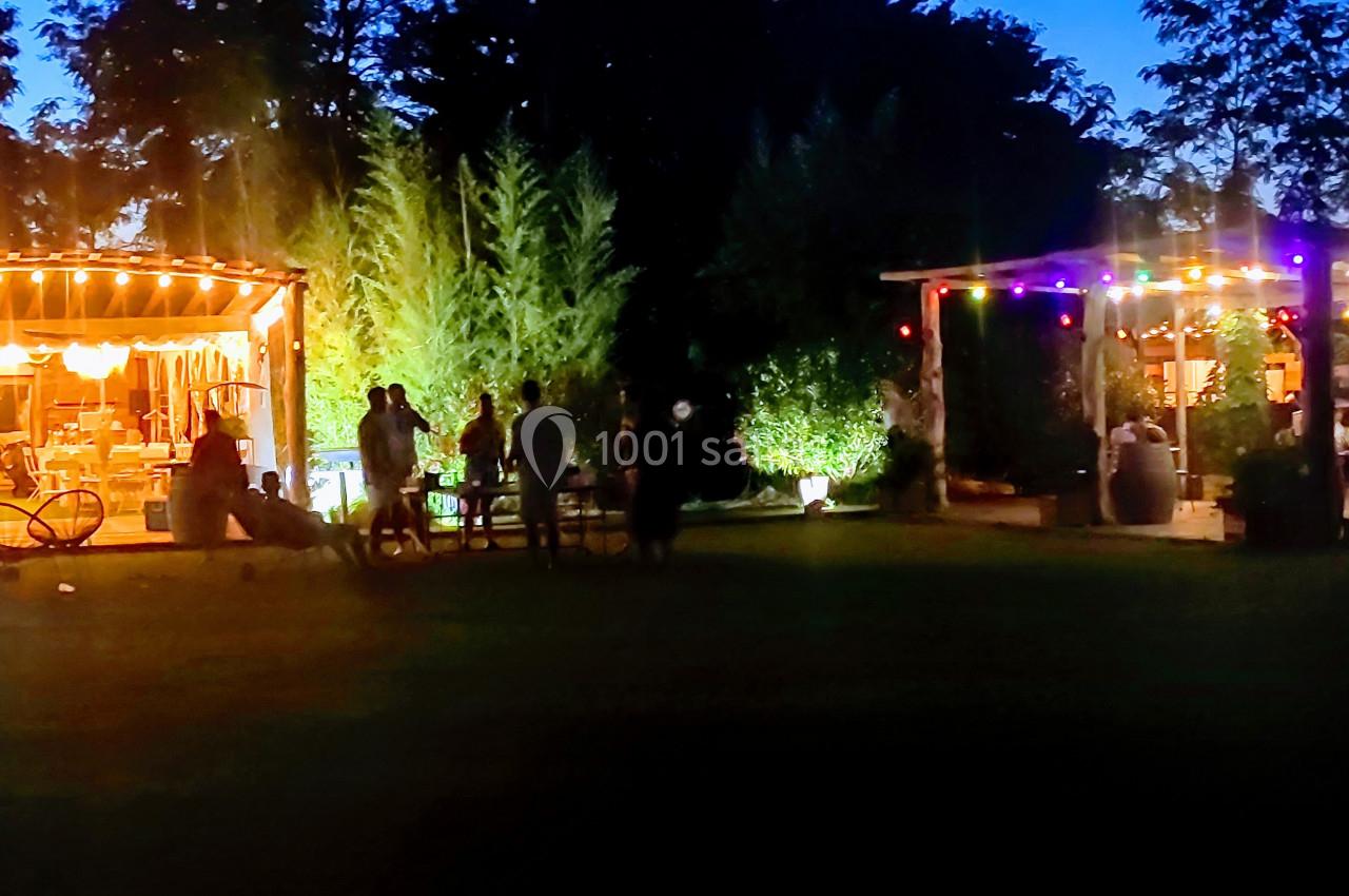 Deux terrasses éclairées par des guirlandes lumineuses dans un jardin en soirée, avec des personnes rassemblées.