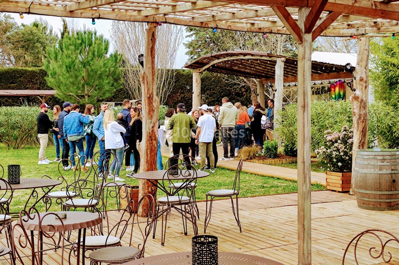 Groupe de personnes rassemblées dans un jardin avec des tables en métal et des barriques en bois au premier plan.