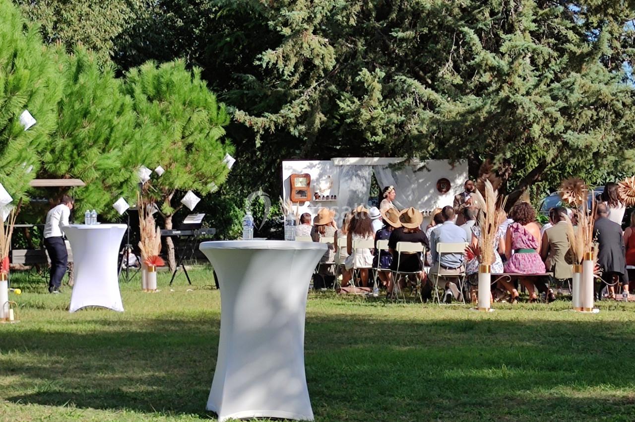 Groupe de personnes assises en extérieur lors d'une cérémonie, entourées d'arbres et de tables hautes décorées.
