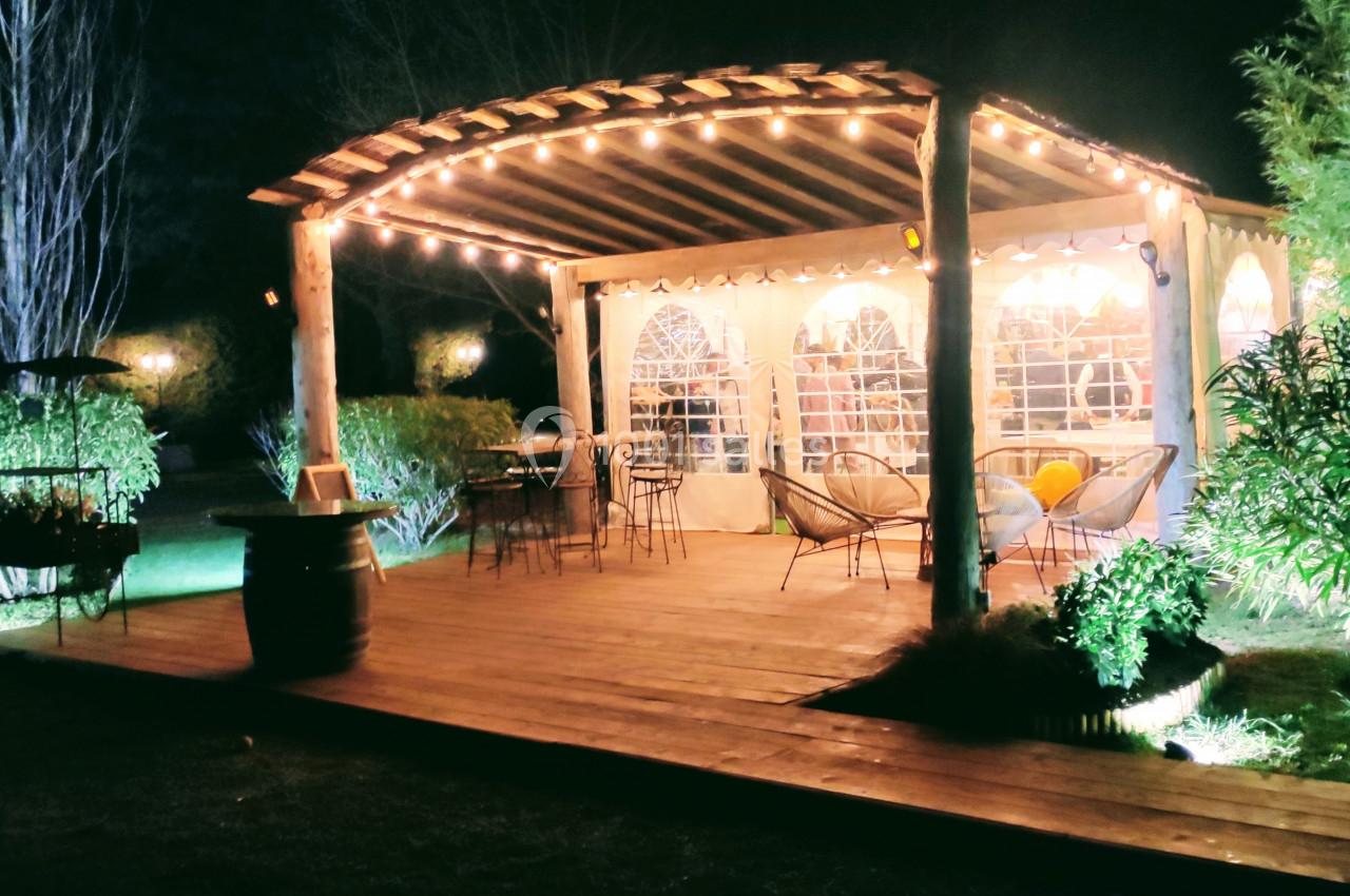 Terrasse en bois éclairée la nuit, avec pergola lumineuse, mobilier de jardin et végétation environnante.