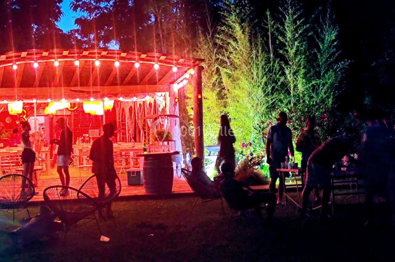 Terrasse éclairée par des guirlandes lumineuses, entourée de végétation, avec des silhouettes de personnes en soirée.