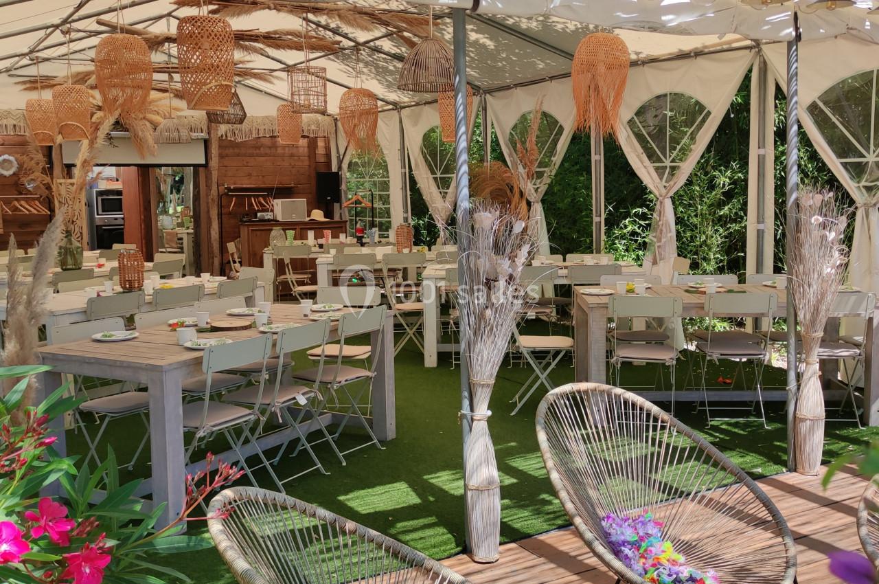 Salle extérieure aménagée sous une tente avec des tables, chaises pliantes et décorations en rotin, entourée de verdure.