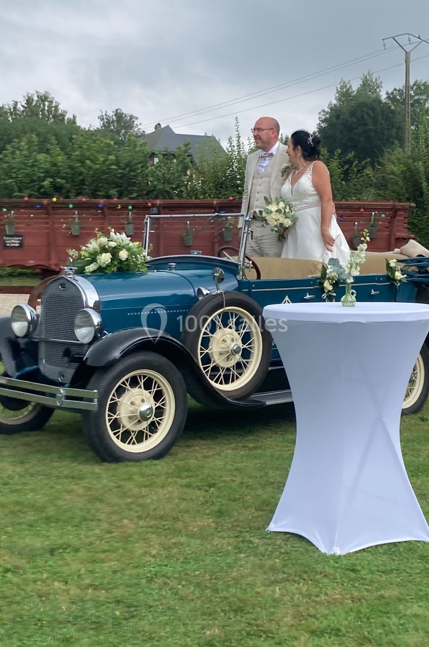 Un couple en tenue de mariage pose près d'une voiture ancienne bleue décorée de fleurs, sur une pelouse.
