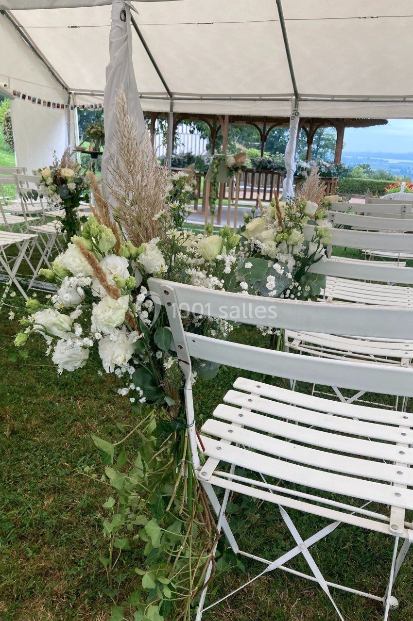 Chaises blanches pliantes décorées de fleurs blanches et feuillages sous une tente, dans un cadre extérieur verdoyant.