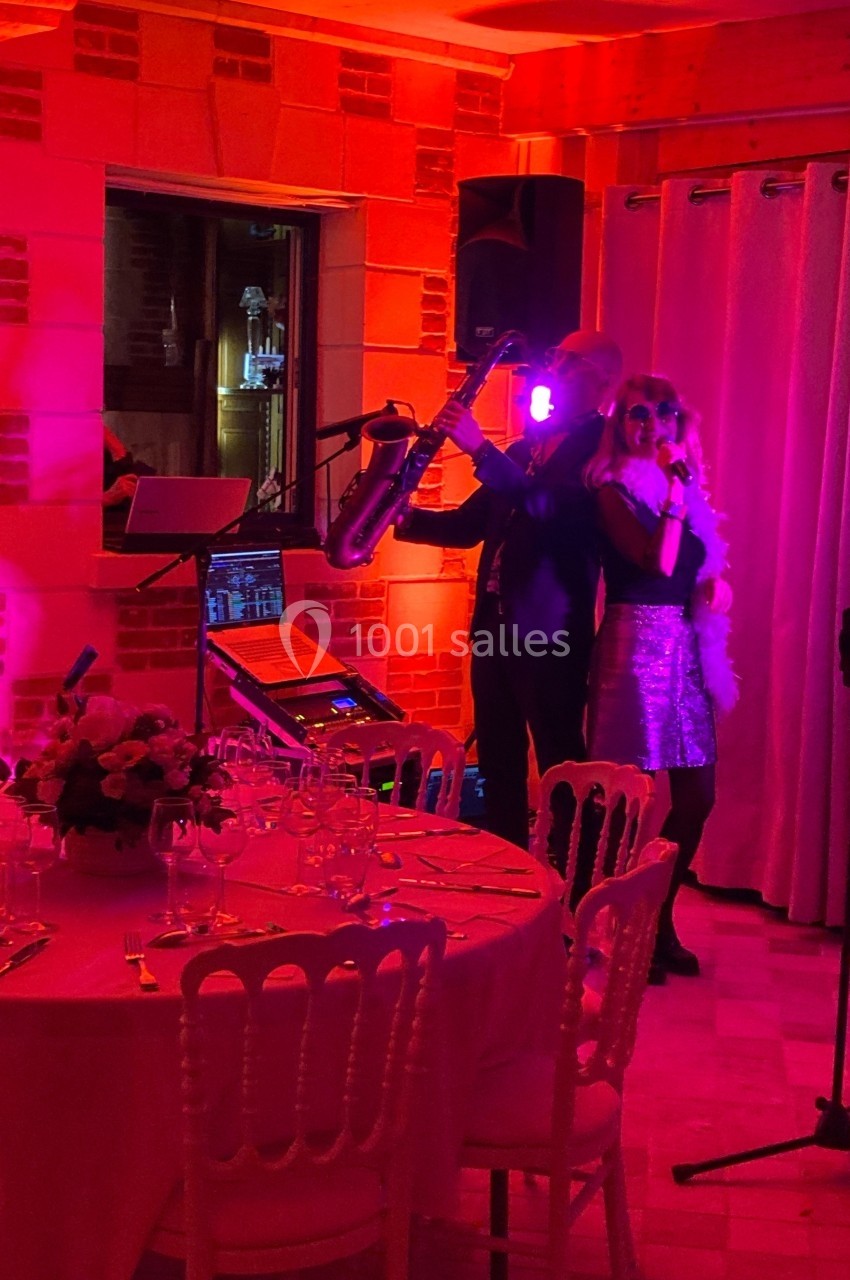 Un duo de musiciens chante et joue du saxophone dans une salle éclairée de lumières roses, près d'une table dressée.