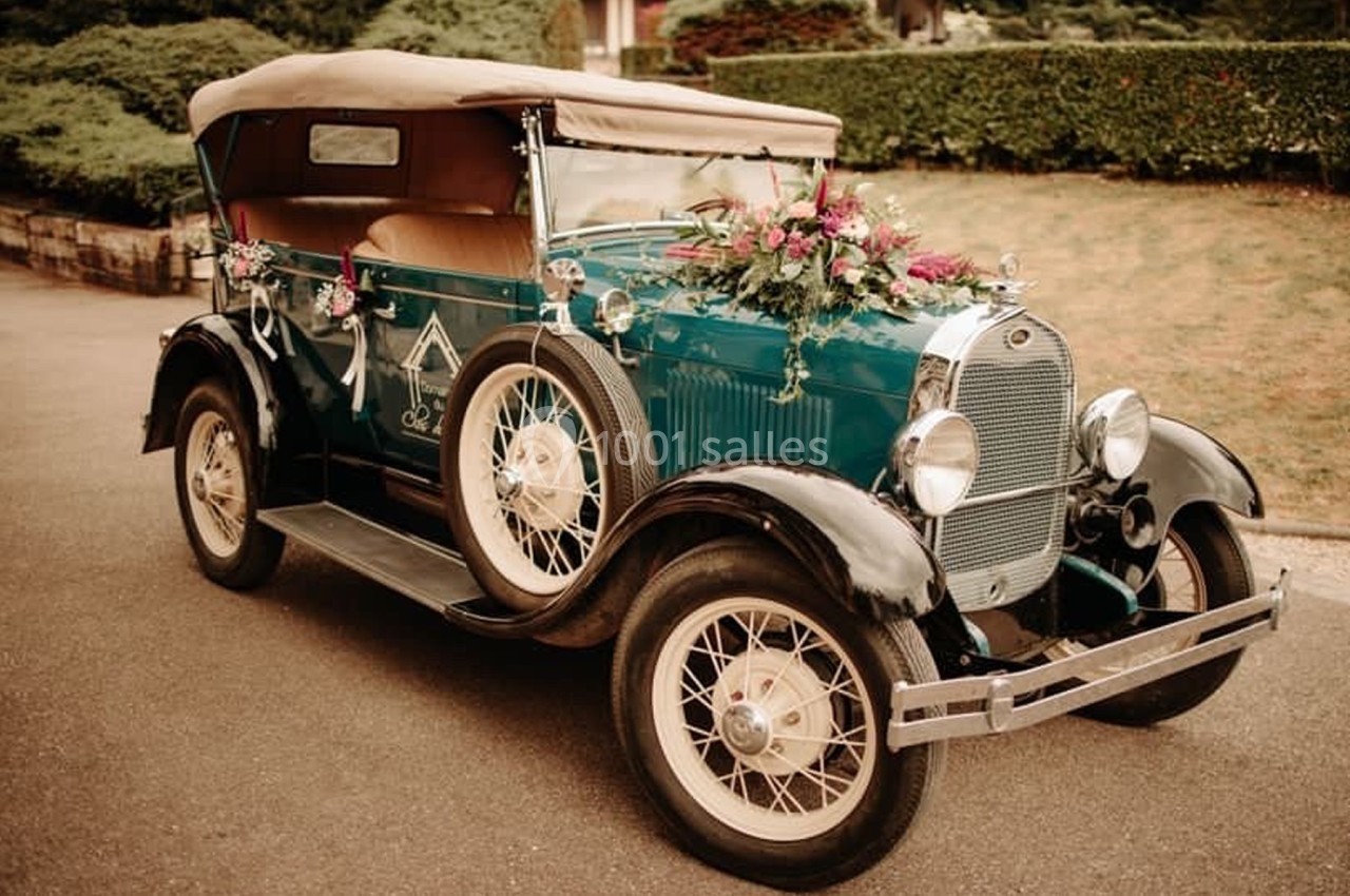 Voiture ancienne décorée de fleurs, stationnée sur une allée pavée devant des haies et un jardin.