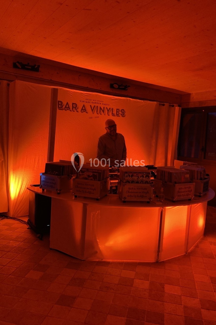 Un DJ mixe derrière un comptoir éclairé en orange, entouré de vinyles, dans une ambiance tamisée.