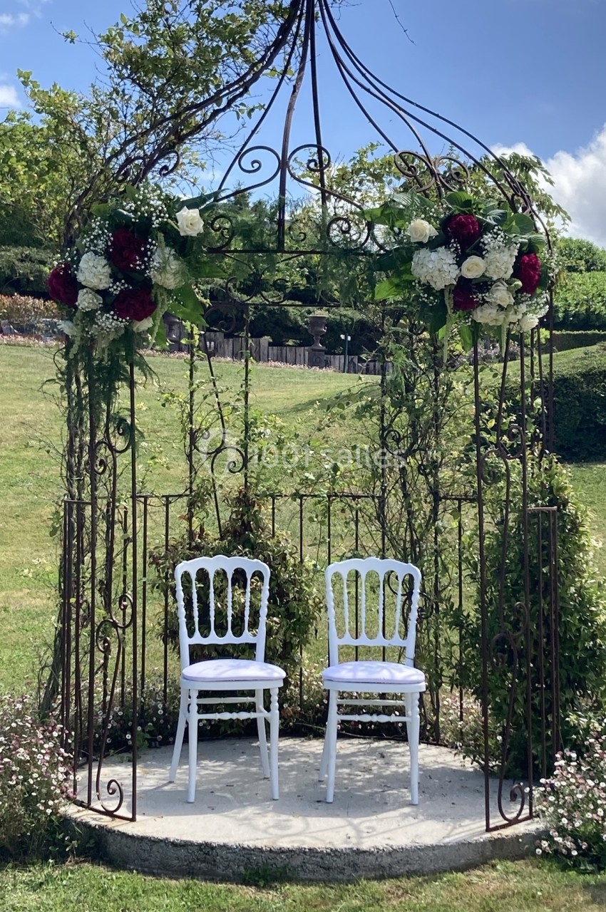 Deux chaises blanches sous une tonnelle en fer forgé décorée de fleurs, dans un jardin verdoyant.