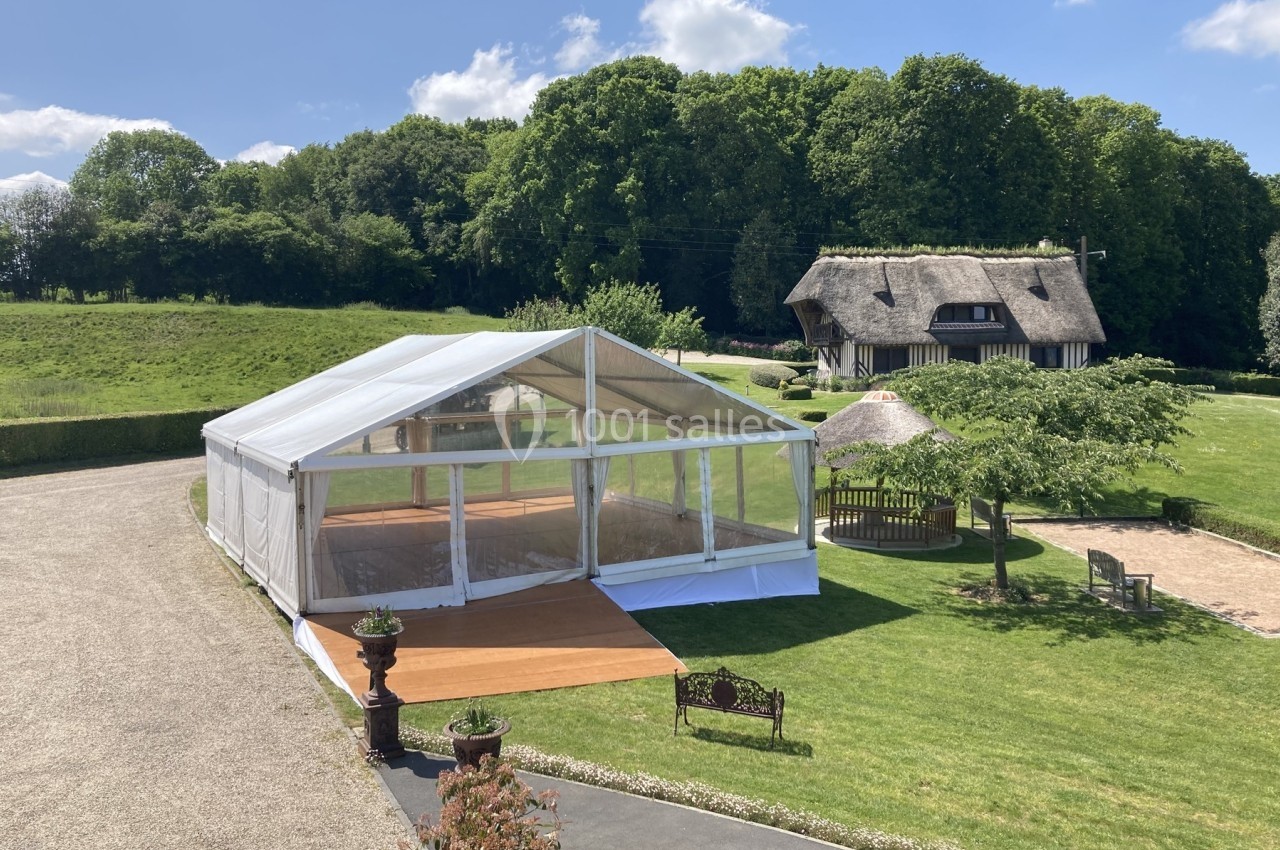 Tente événementielle transparente installée sur une pelouse, entourée de verdure et d'une maison au toit de chaume.