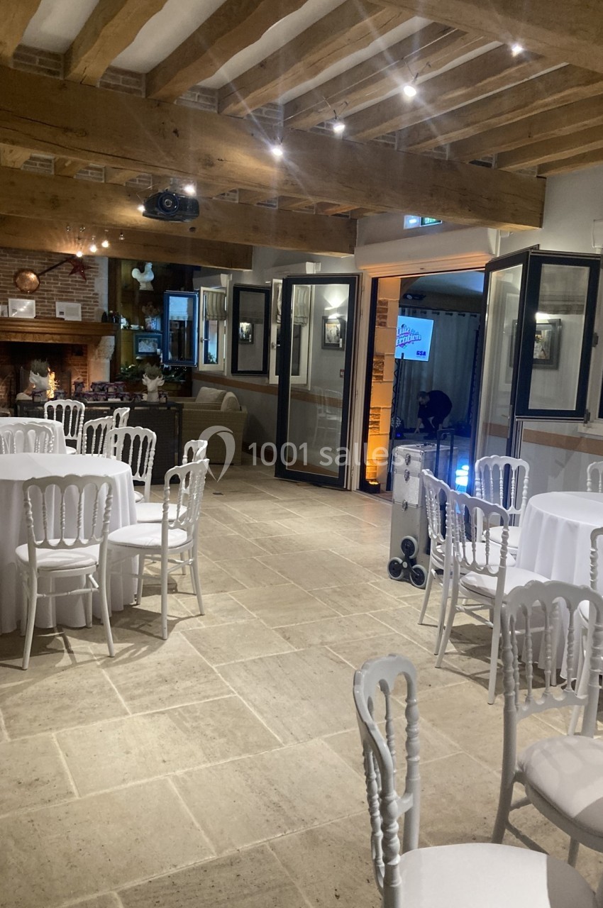 Salle élégante avec tables rondes dressées de nappes blanches, chaises assorties et poutres apparentes au plafond.