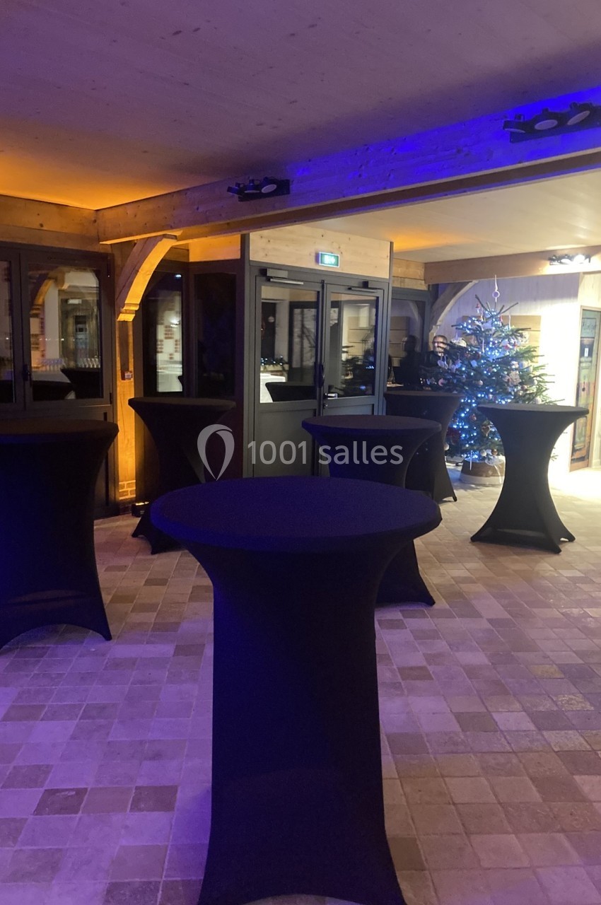 Salle décorée avec des tables hautes noires, un sol en carreaux clairs et un sapin de Noël illuminé en arrière-plan.