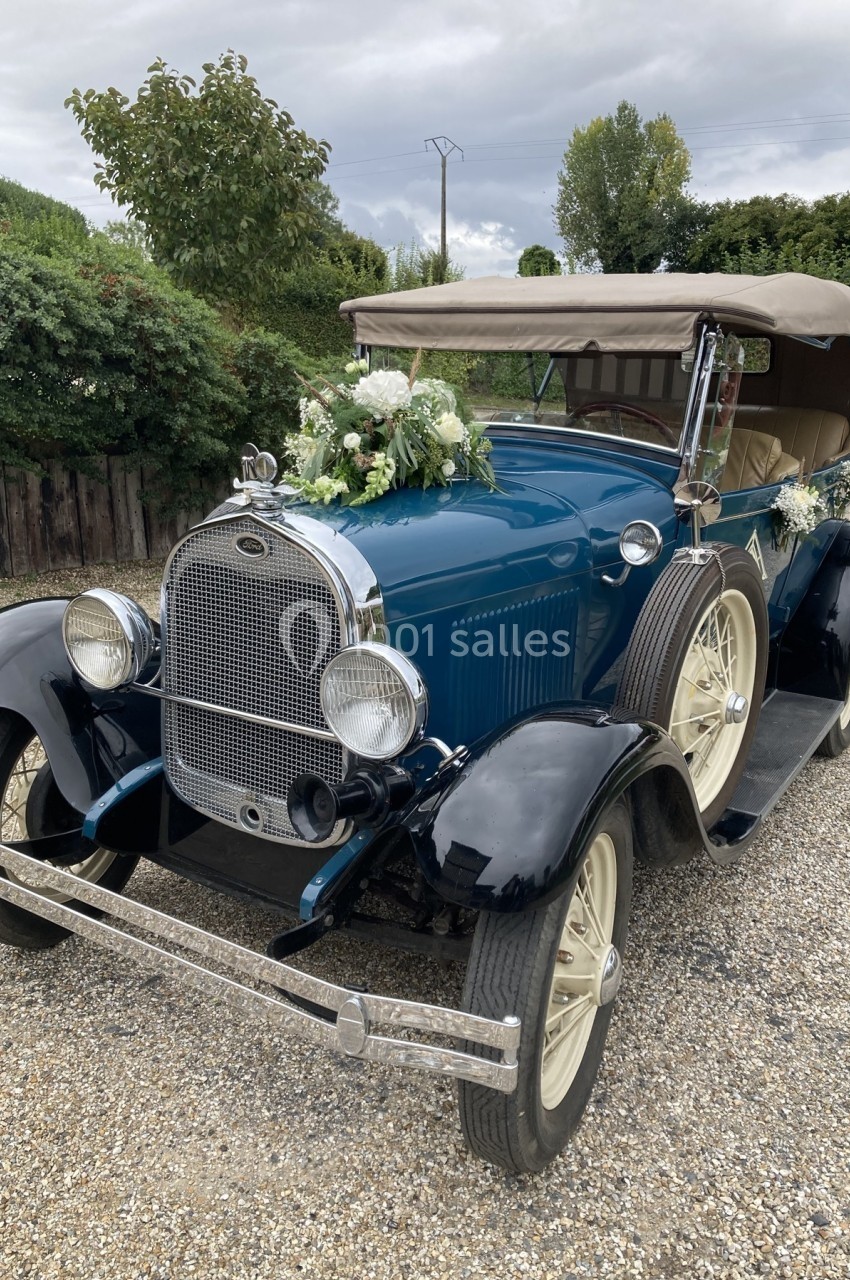 Voiture ancienne bleue avec capote beige, ornée de fleurs blanches, stationnée sur une allée gravillonnée.