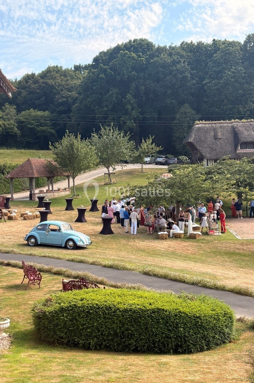 Groupe de personnes rassemblées en extérieur près de tables hautes, avec une voiture bleue et un paysage champêtre en…