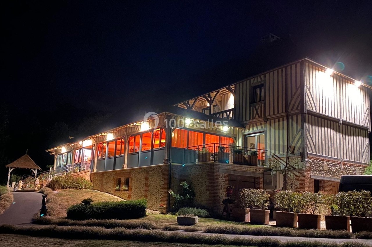 Bâtiment à colombages éclairé la nuit, avec une terrasse vitrée illuminée et un jardin environnant.