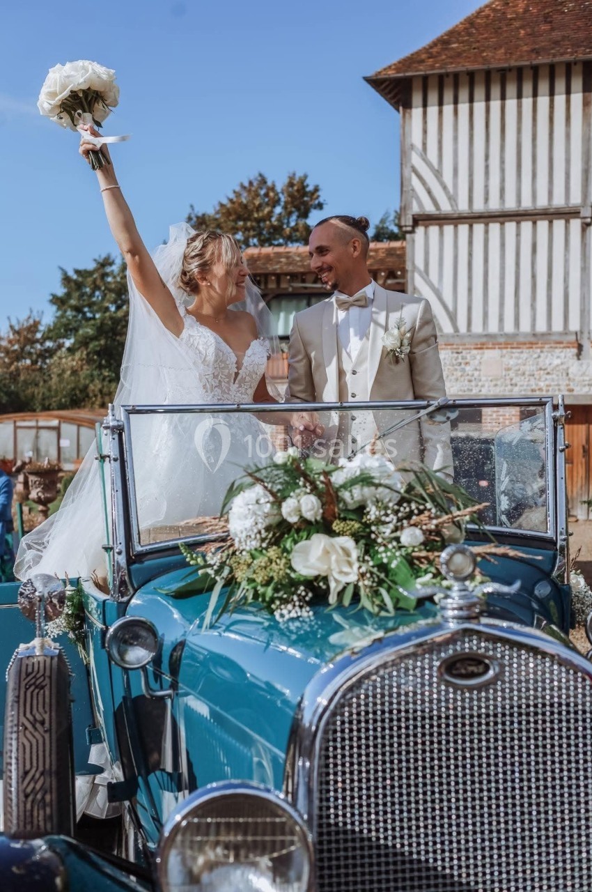 Un couple de mariés souriants dans une voiture ancienne décorée de fleurs, la mariée levant un bouquet.