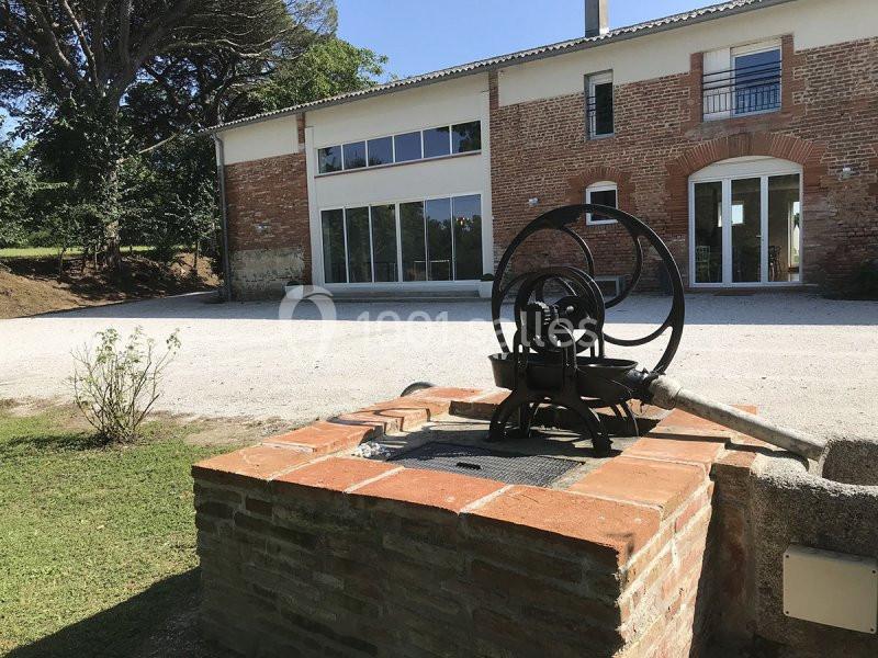 Pompe à eau ancienne en métal sur un puits en briques, devant une maison en briques avec de grandes fenêtres.