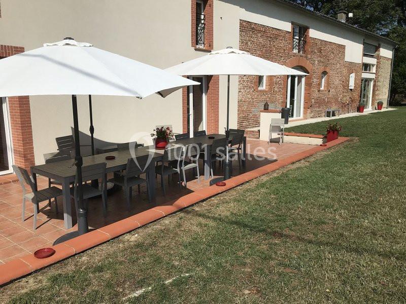 Terrasse avec tables, chaises en bois gris et parasols blancs, devant une maison en briques et crépi beige.