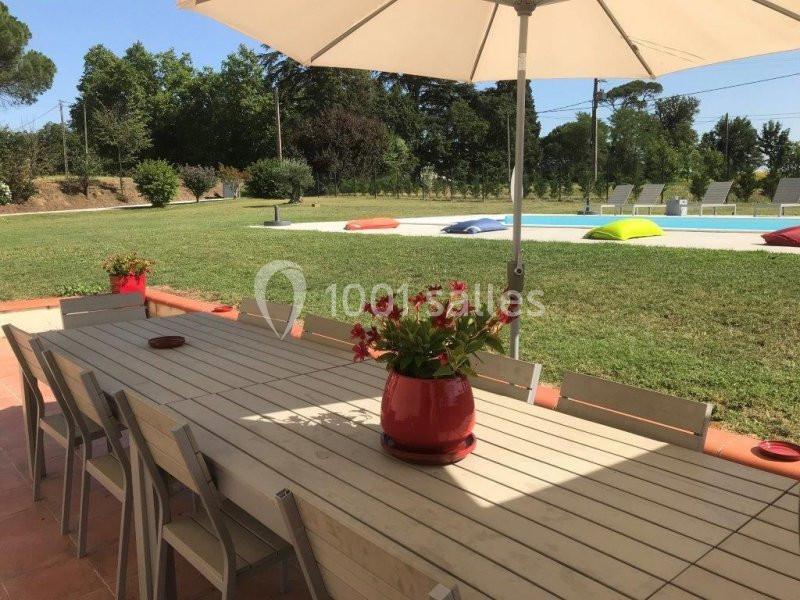 Table en bois avec des chaises, un pot de fleurs rouges, sous un parasol, près d'une pelouse et d'une piscine.