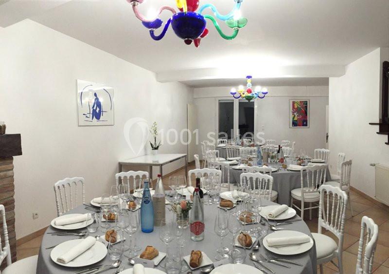 Salle de réception décorée avec des tables rondes dressées, nappes grises, chaises blanches et lustres colorés.