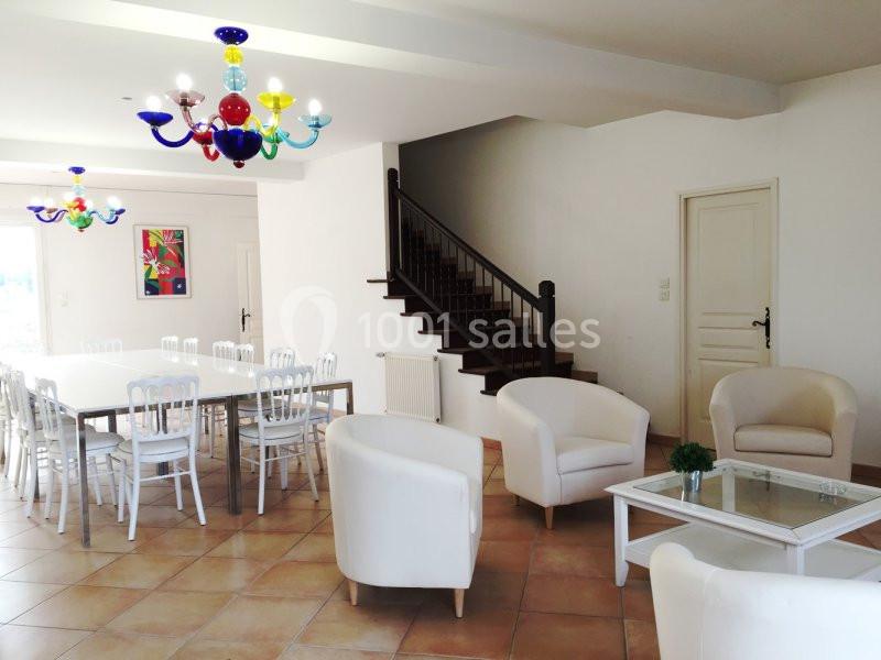 Salle lumineuse avec tables blanches, chaises, fauteuils, escaliers en bois, et lustres colorés.