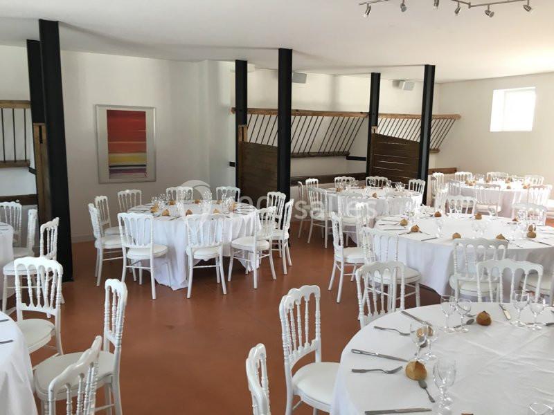 Salle de réception avec tables rondes dressées de nappes blanches, chaises en bois blanc et lumière naturelle.