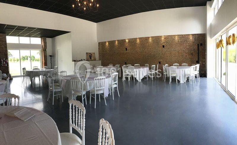 Salle lumineuse avec tables rondes dressées, chaises blanches et murs en briques, éclairée par des baies vitrées.