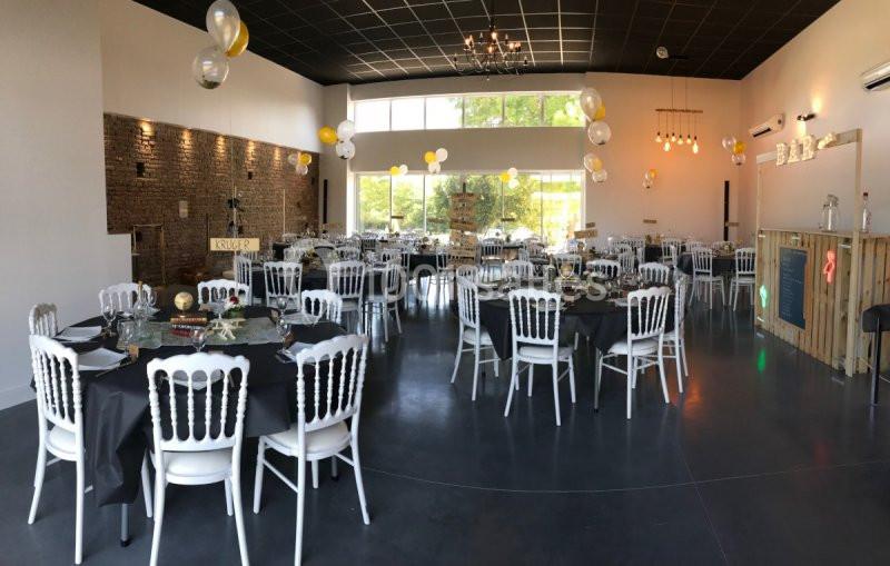 Salle de réception décorée avec des tables rondes, nappes noires, chaises blanches et ballons suspendus.