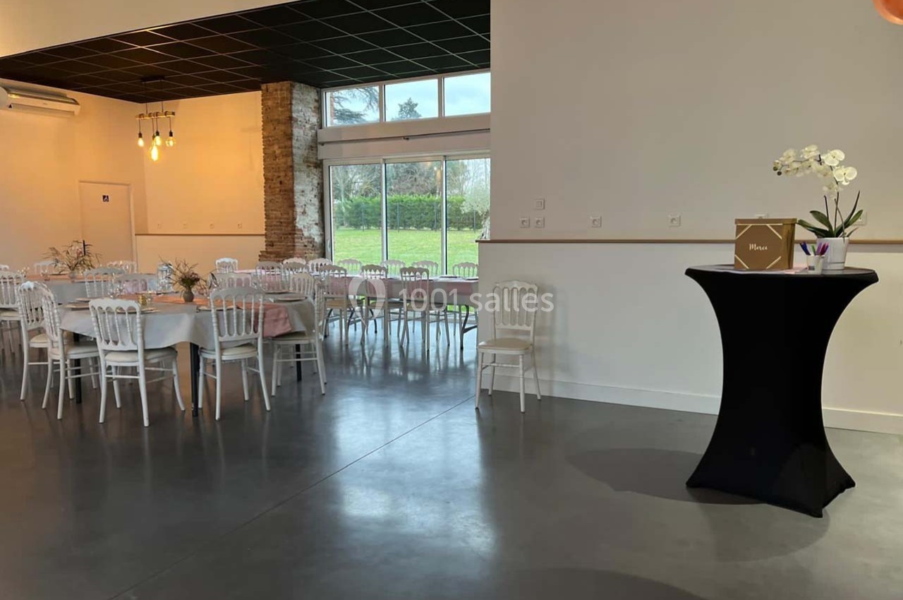Salle de réception lumineuse avec tables dressées, chaises blanches, grandes baies vitrées et un pupitre décoré.