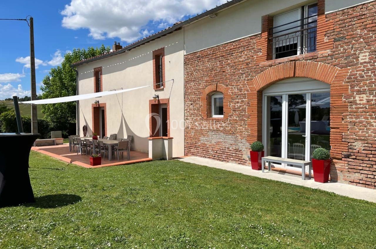 Façade d'une maison en briques et crépi avec terrasse aménagée, pelouse et ciel dégagé en arrière-plan.