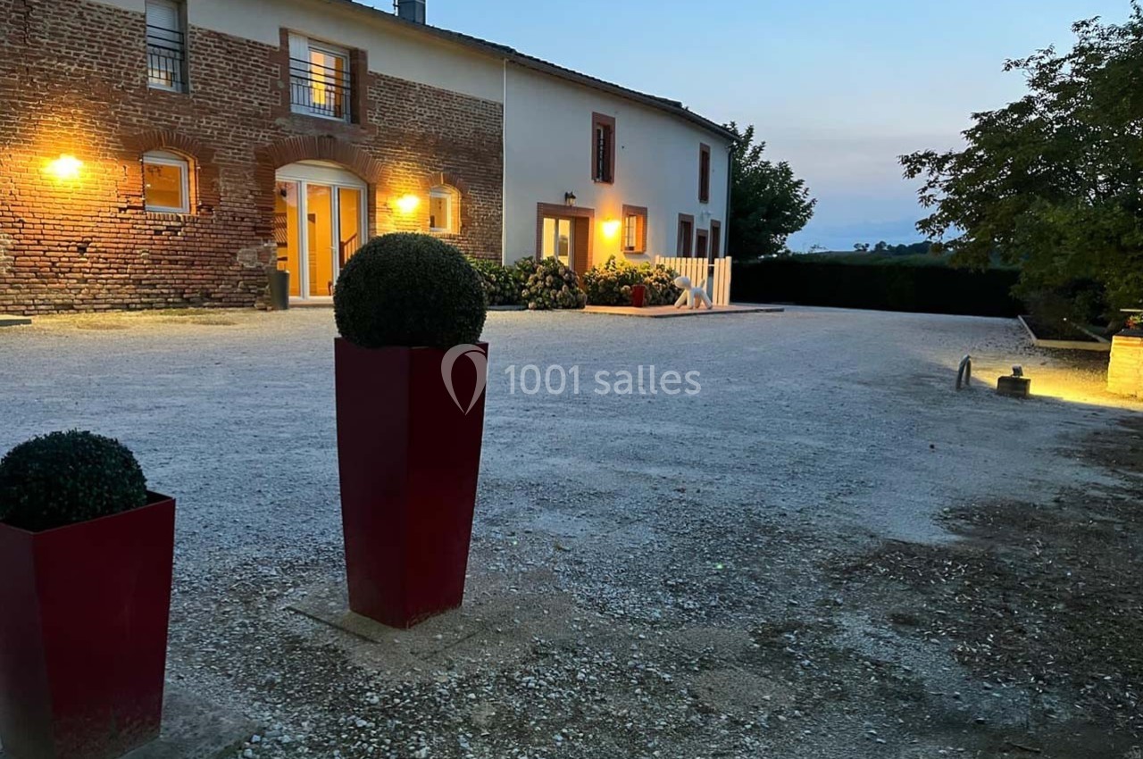 Cour gravillonnée devant une maison en briques et crépi, éclairée par des lumières extérieures au crépuscule.