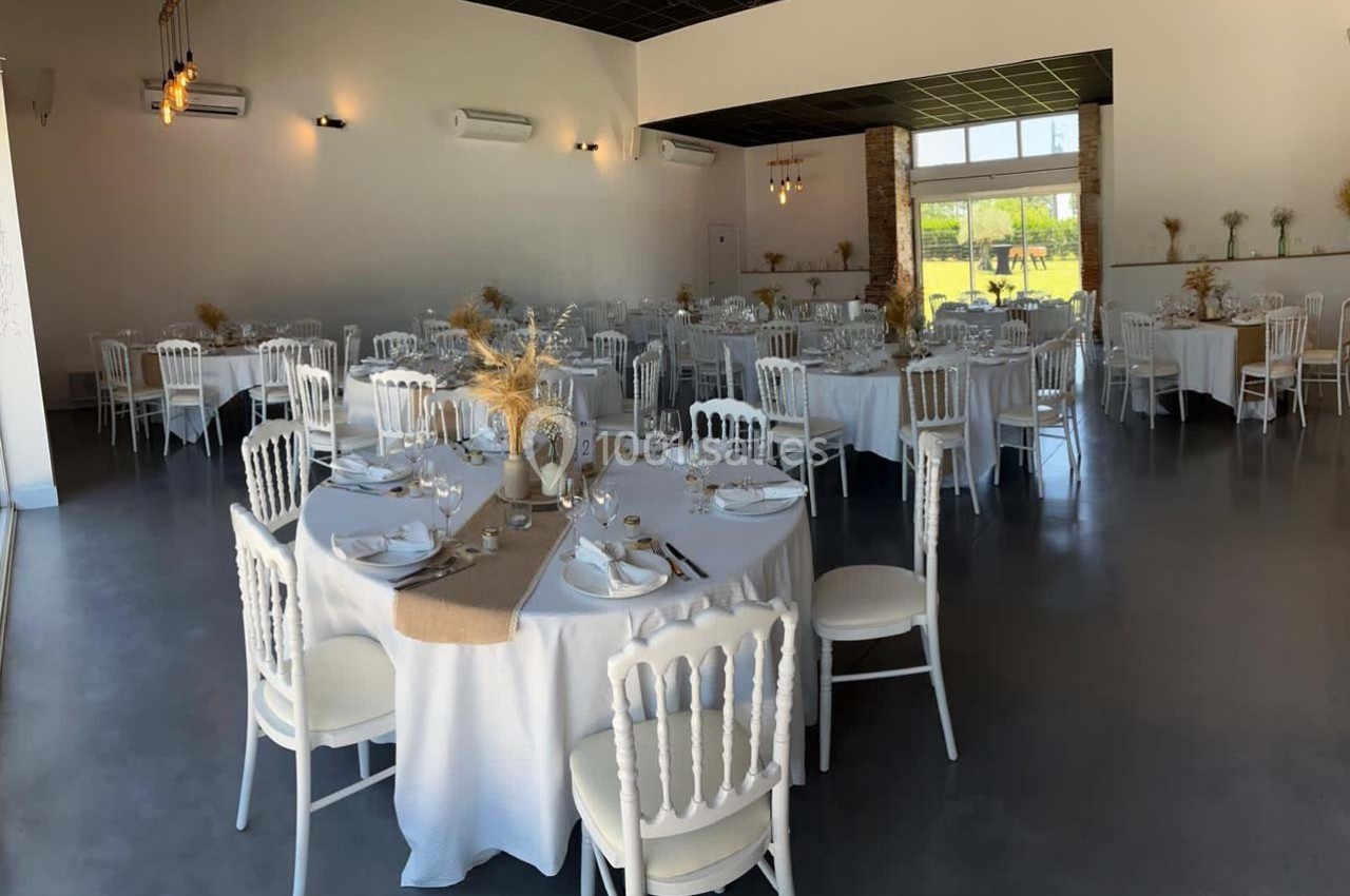 Salle de réception lumineuse avec tables rondes dressées, nappes blanches et décorations florales naturelles.