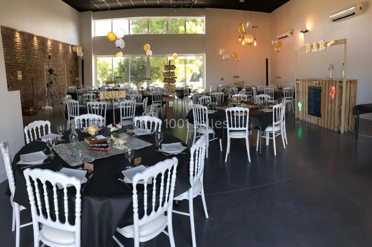 Salle décorée pour un événement, avec tables rondes dressées, chaises blanches et guirlandes lumineuses suspendues.