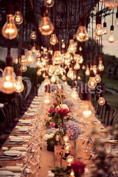 Table longue décorée avec des fleurs et des verres, éclairée par de nombreuses ampoules suspendues dans un cadre extérieur.