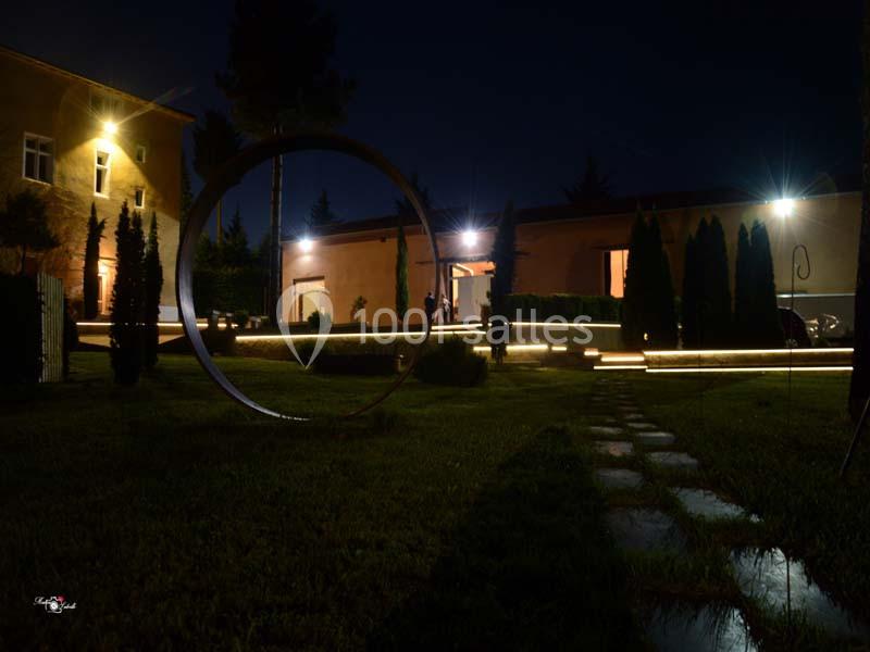 Cour éclairée la nuit avec un anneau décoratif au premier plan, des arbres et des bâtiments en arrière-plan.
