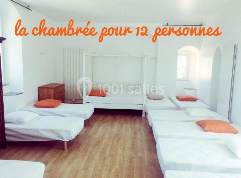 Dortoir lumineux avec lits simples alignés, draps blancs et quelques oreillers colorés, sol en parquet.