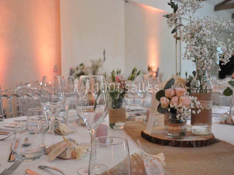 Table décorée pour un mariage avec nappes blanches, verres, fleurs pastel et bougies dans une salle éclairée.