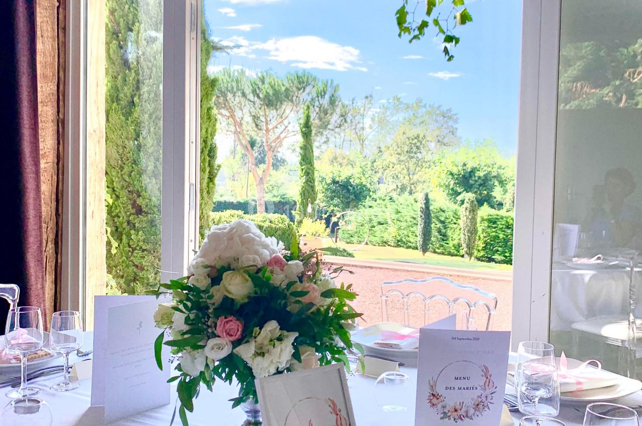 Table élégamment dressée avec des fleurs et des menus, donnant sur un jardin verdoyant visible à travers une baie vitrée.