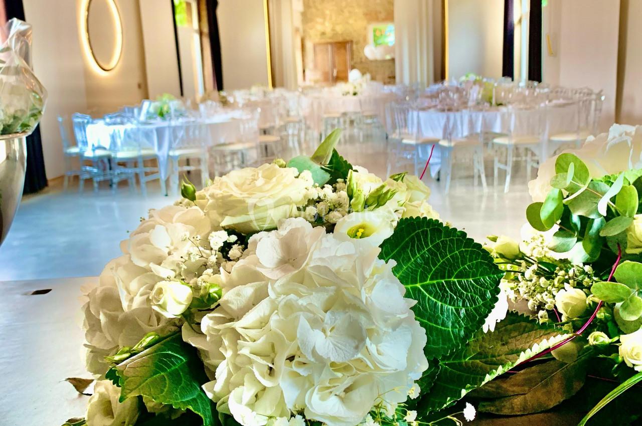 Salle de réception lumineuse décorée pour un événement, avec des bouquets de fleurs blanches au premier plan.