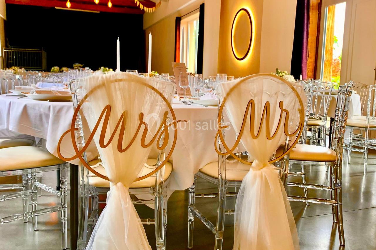 Chaises décorées avec les inscriptions ’Mrs’ et ’Mr’, entourées de tables dressées dans une salle de réception lumineuse.