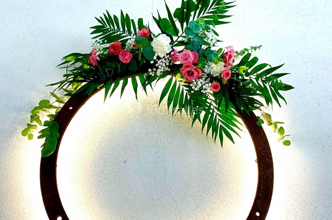 Couronne lumineuse circulaire décorée de feuillage vert et de fleurs roses et blanches, accrochée à un mur clair.