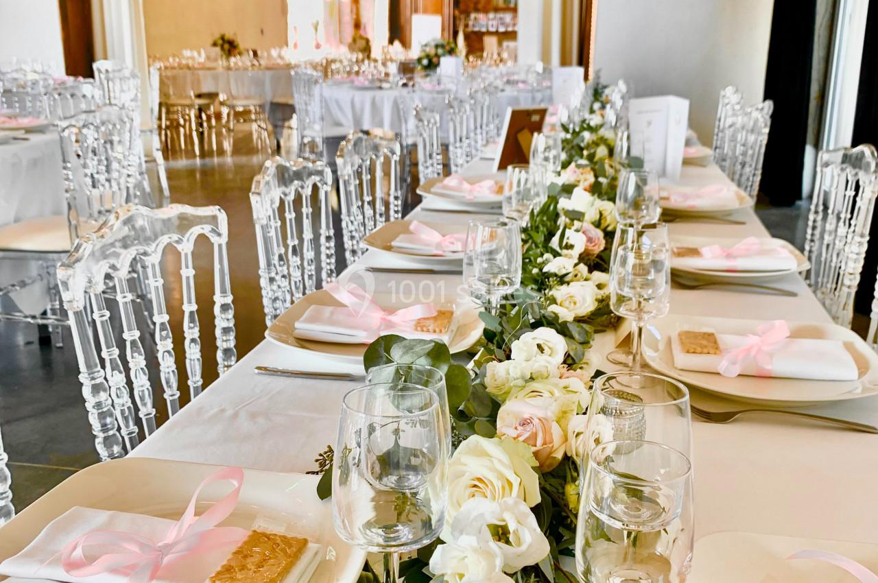 Table décorée pour un événement avec nappes blanches, fleurs, rubans roses et chaises transparentes dans une salle lumineuse.