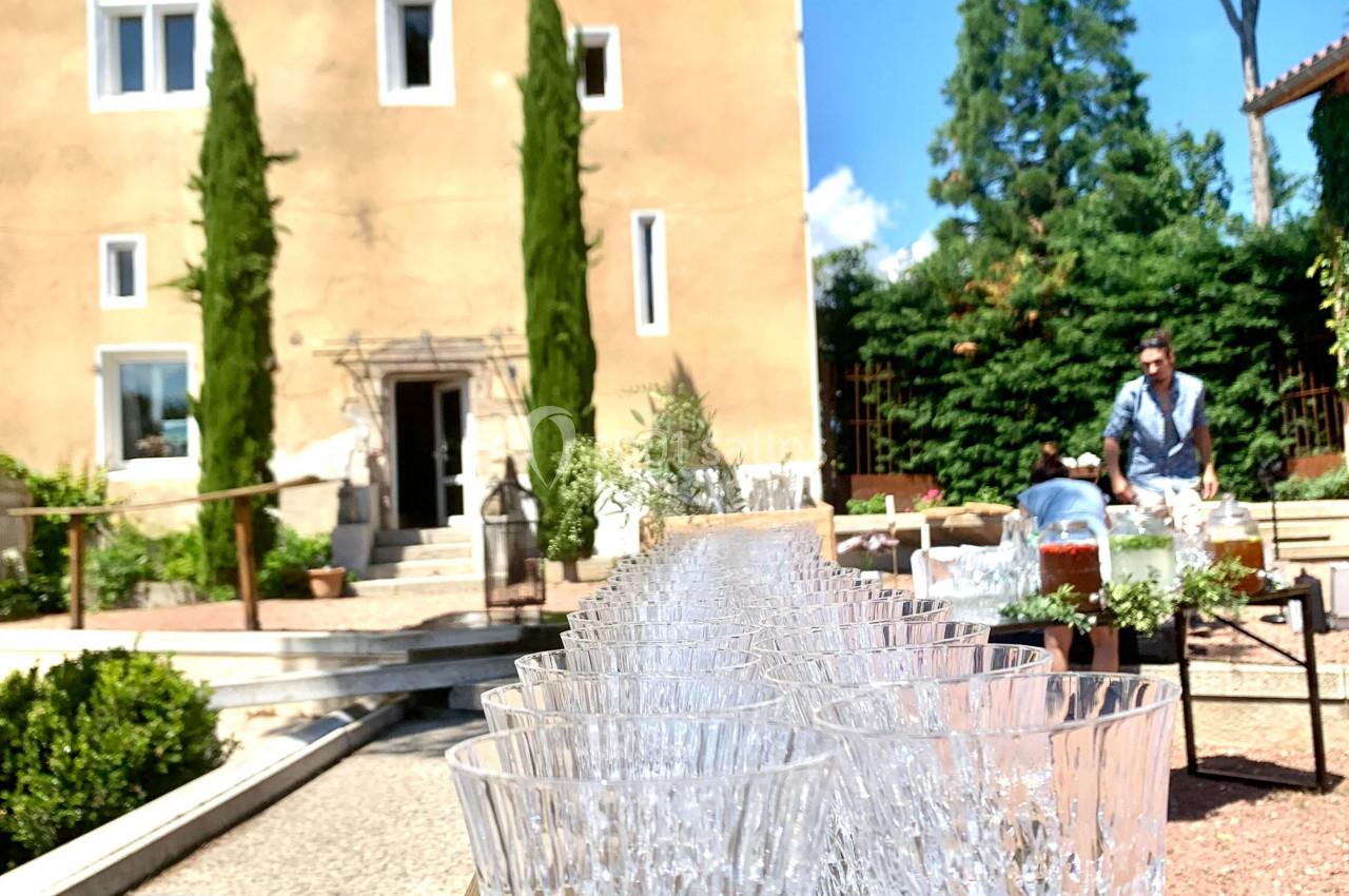 Ligne de verres en cristal sur une table en bois devant une maison ancienne entourée de verdure.